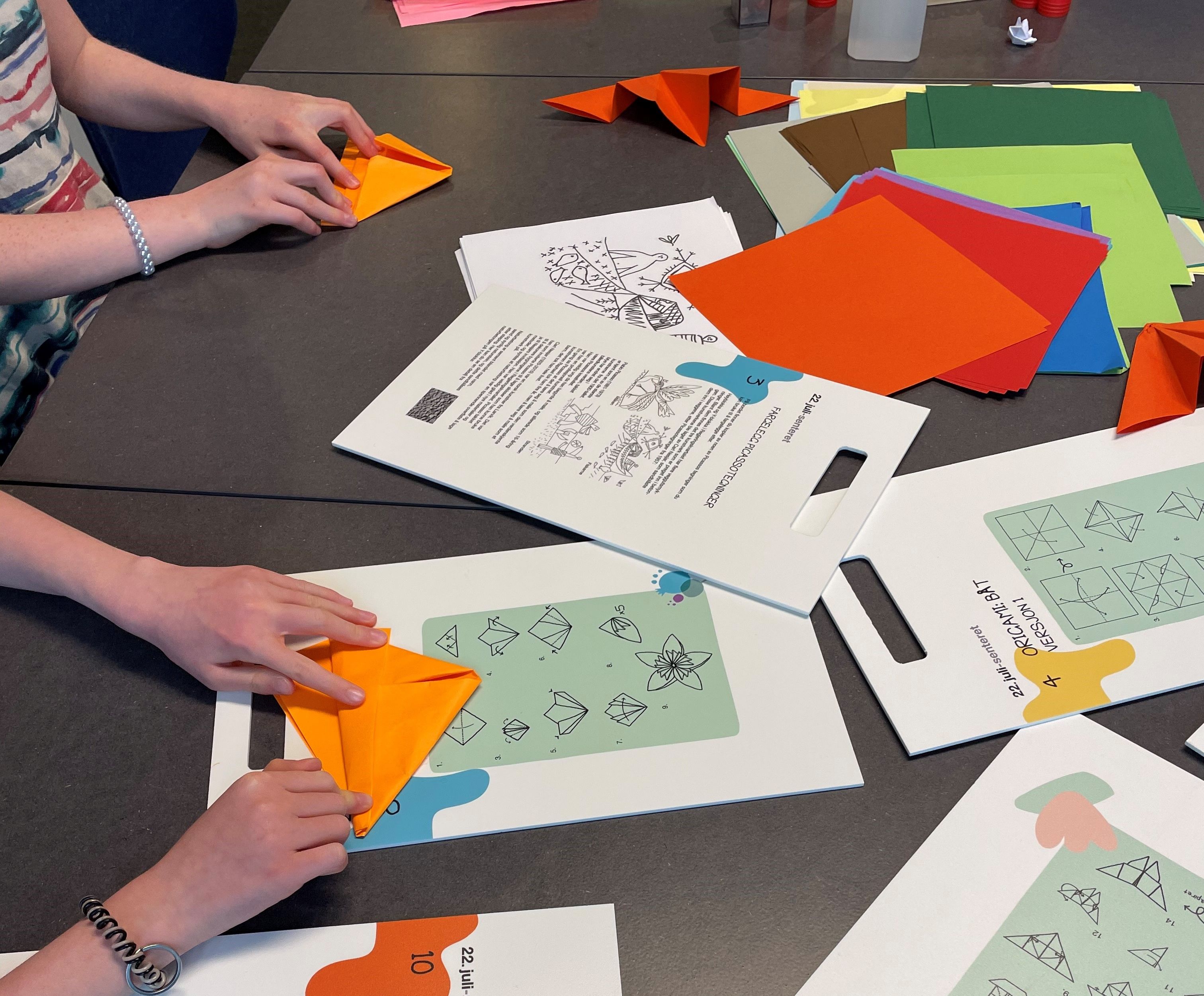 Bord med flere hender som bretter origami figurer. Oppskrifter ligger på bordet. Fargerike ark.