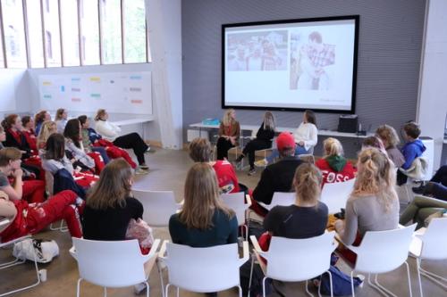 Three women sit in chairs. An audience watches them. Two pictures of people are shown on a large screen.