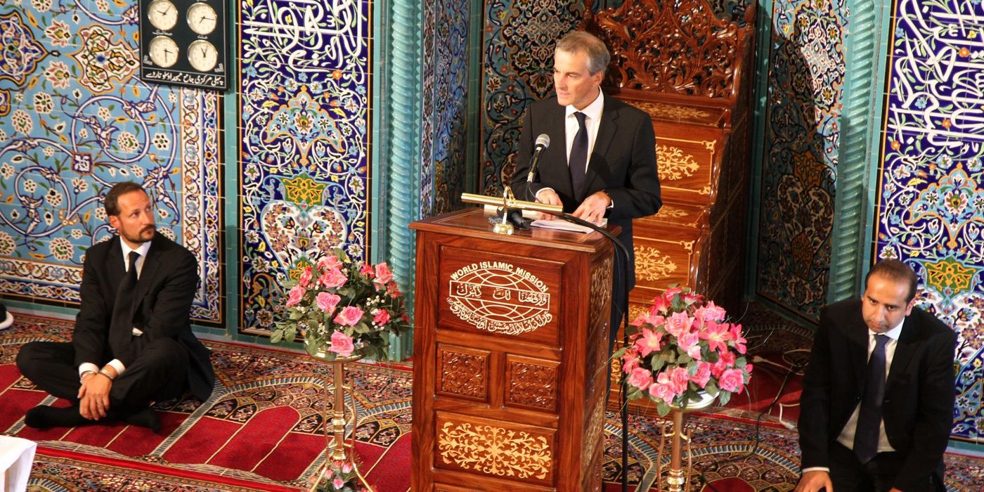One in a suit on a pedestal, two men in suits sitting on the floor. Ornate wall in blue behind. Flowers and red carpet on the floor.