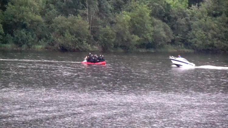 Screen shot from video recording. Water in the foreground. A red boat carrying unknown number of people. A white boat carrying one person. Trees and bushes in the background. 