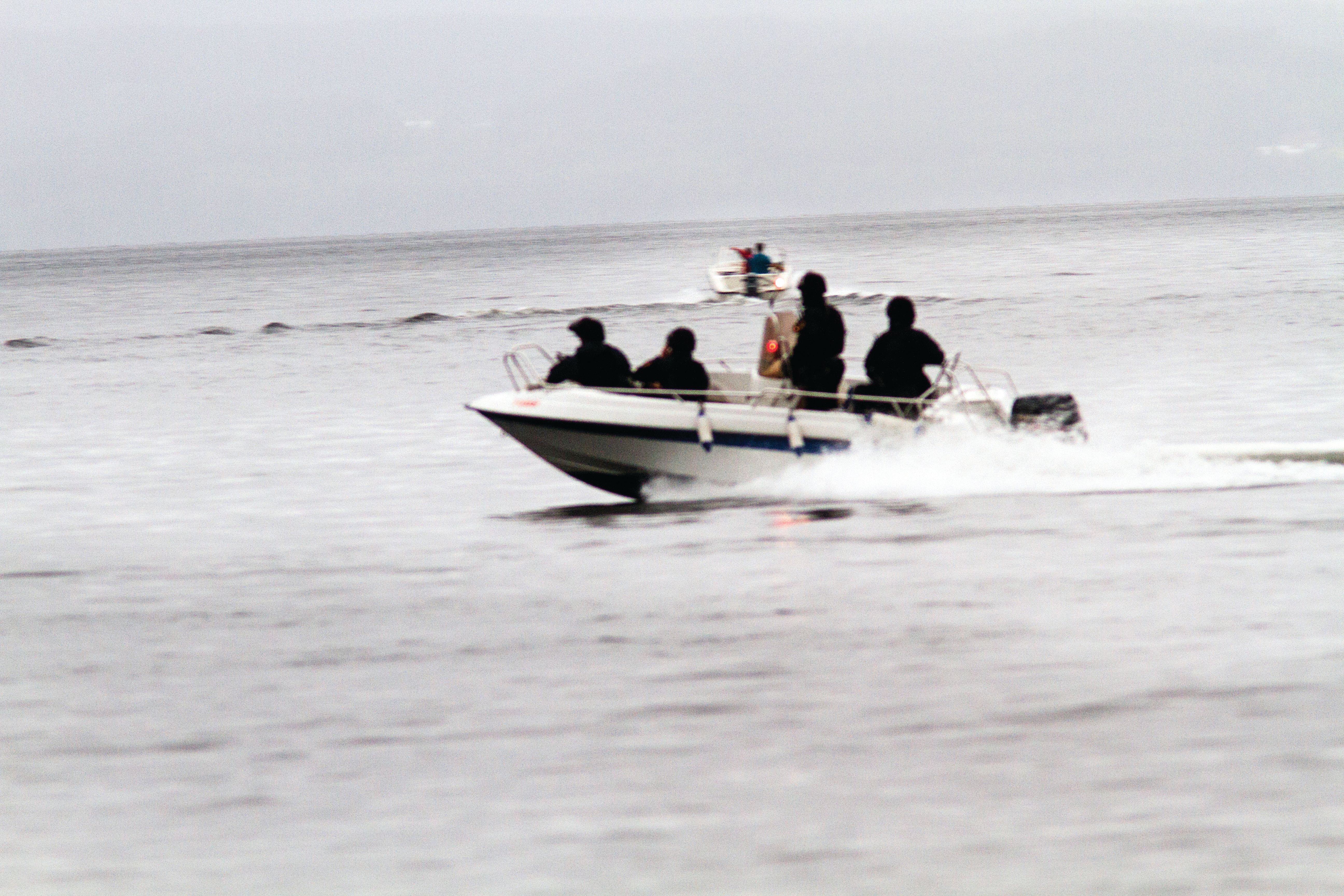 Vann. En båt med fire mennesker i uniform i forkant. En båt i bakgrunnen.