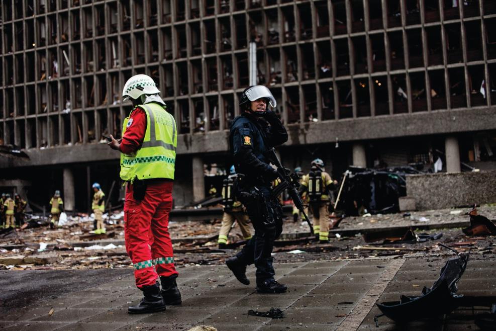 en brannmann og et bevæpnet politi foran en ødelagt bygning. Flere innsatsstyrker i bakgrunnen. Store fysiske ødeleggelser.
