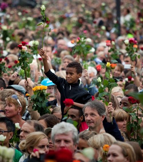 Menneskemengde samlet. Voksne og barn med roser i hendene. I midten en gutt som sitter på skuldrene til en mann, mens han løfter en hvit rose i høyre hånd.
