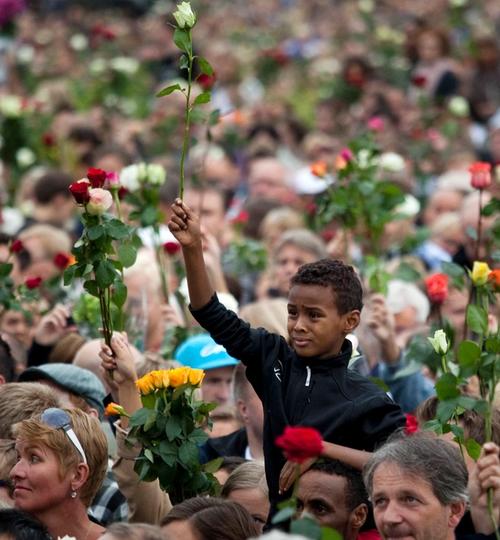 Many people gathered in a crowd. Adults and children carrying roses. In the centre, a boy lifting a white rose, sitting on a mans shoulders.