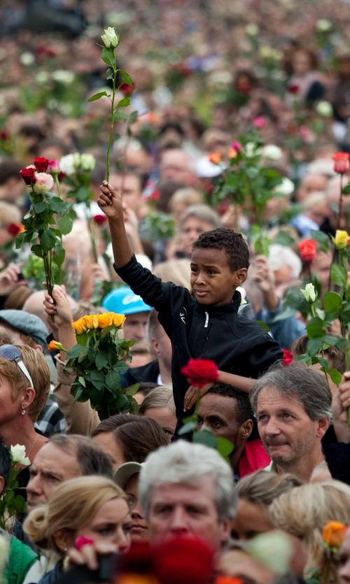 Menneskemengde samlet. Voksne og barn med roser i hendene. I midten en gutt som sitter på skuldrene til en mann, mens han løfter en hvit rose i høyre hånd.