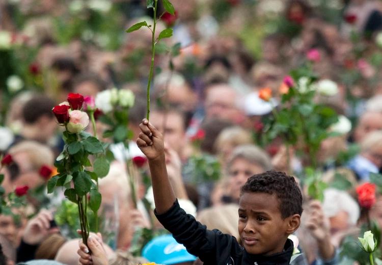 Many people gathered in a crowd. Adults and children carrying roses. In the centre, a boy lifting a white rose, sitting on a mans shoulders.