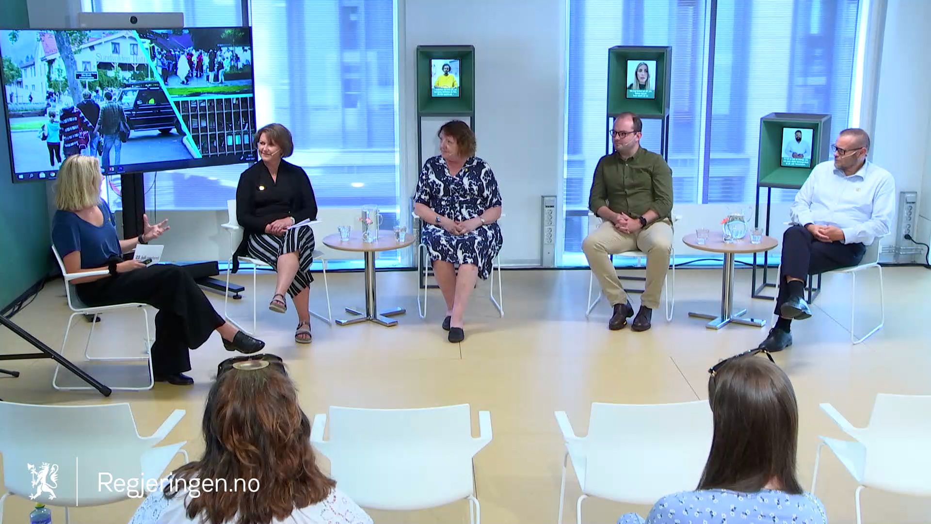 Five people sit in a panel discussion in front of a screen showing blurry images of people and buildings. three smaller screens in stands are seen behind the panelists. The audience sits in front.