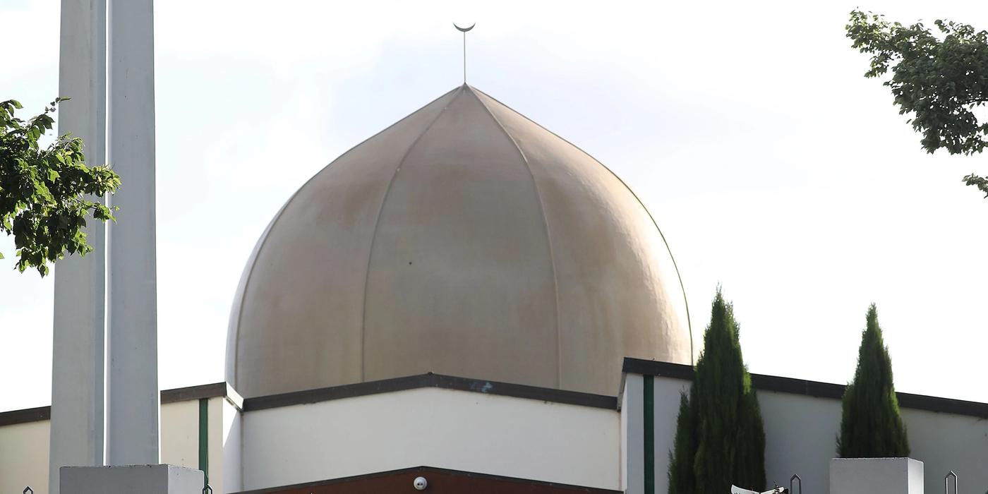 Red barrier in front of a crowd. Mosque in white with a tall tower. Brown/grey dome.