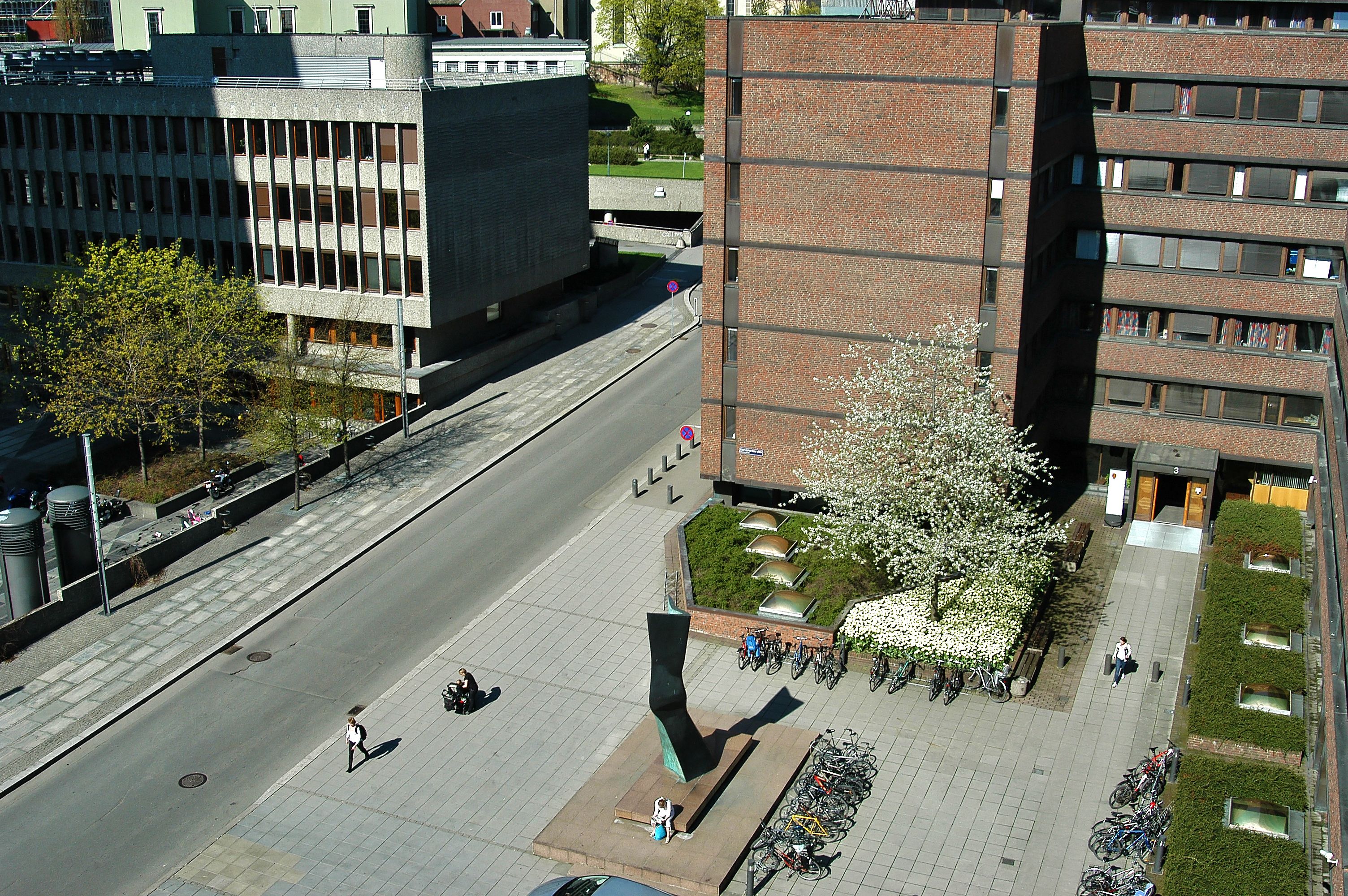 A square with some people. An abstract statue of green metal in the middle of the square. Some green plants and a road.