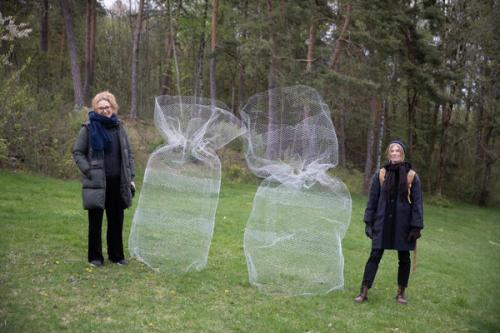 Two warmly dressed women stand on a lawn next to two wire sculptures that resemble large sacks. Trees in the background. Photo