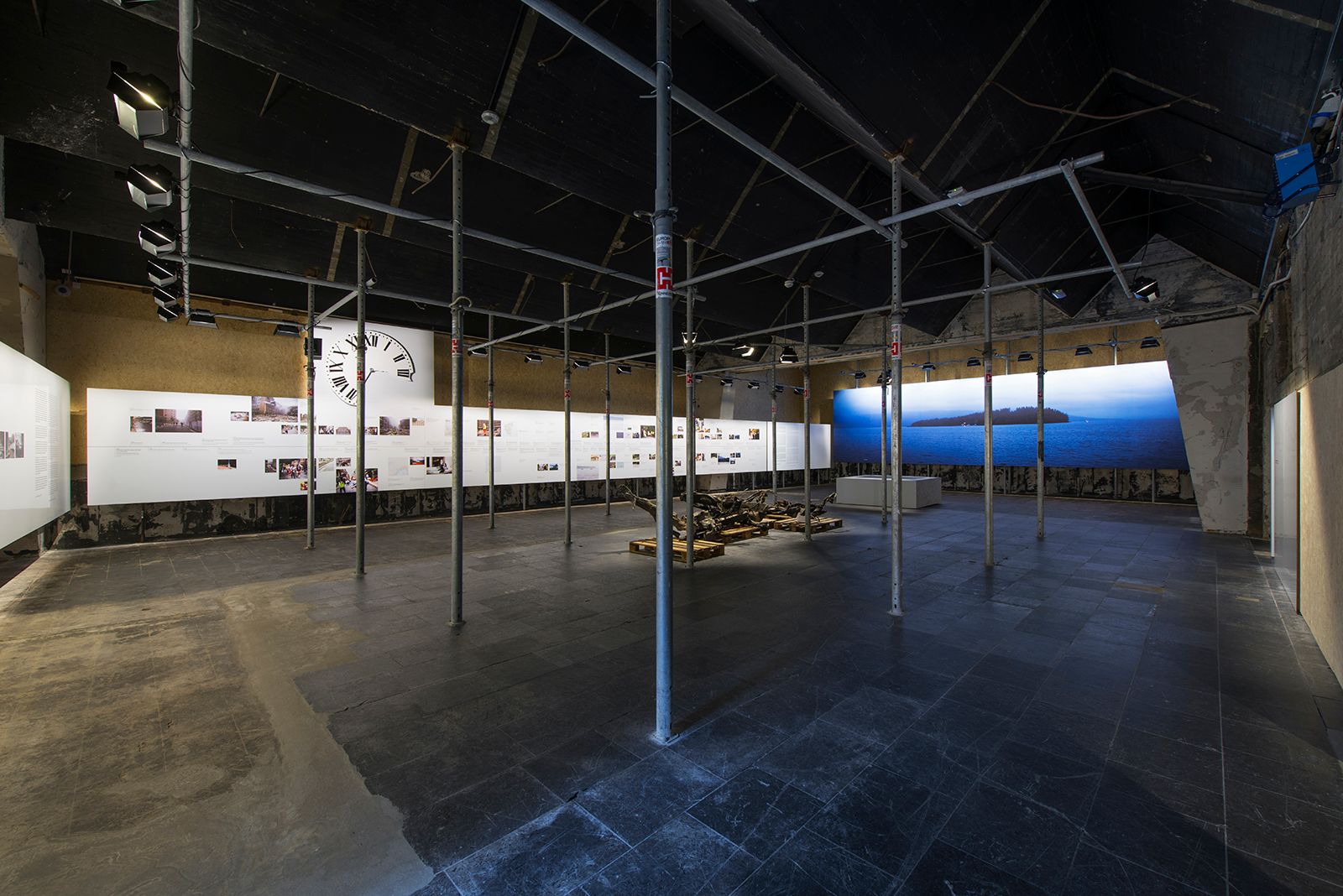  Dark room with illuminated exhibition walls with text and images. A landscape photograph of water and forest. Steel framework, and a car wreck.