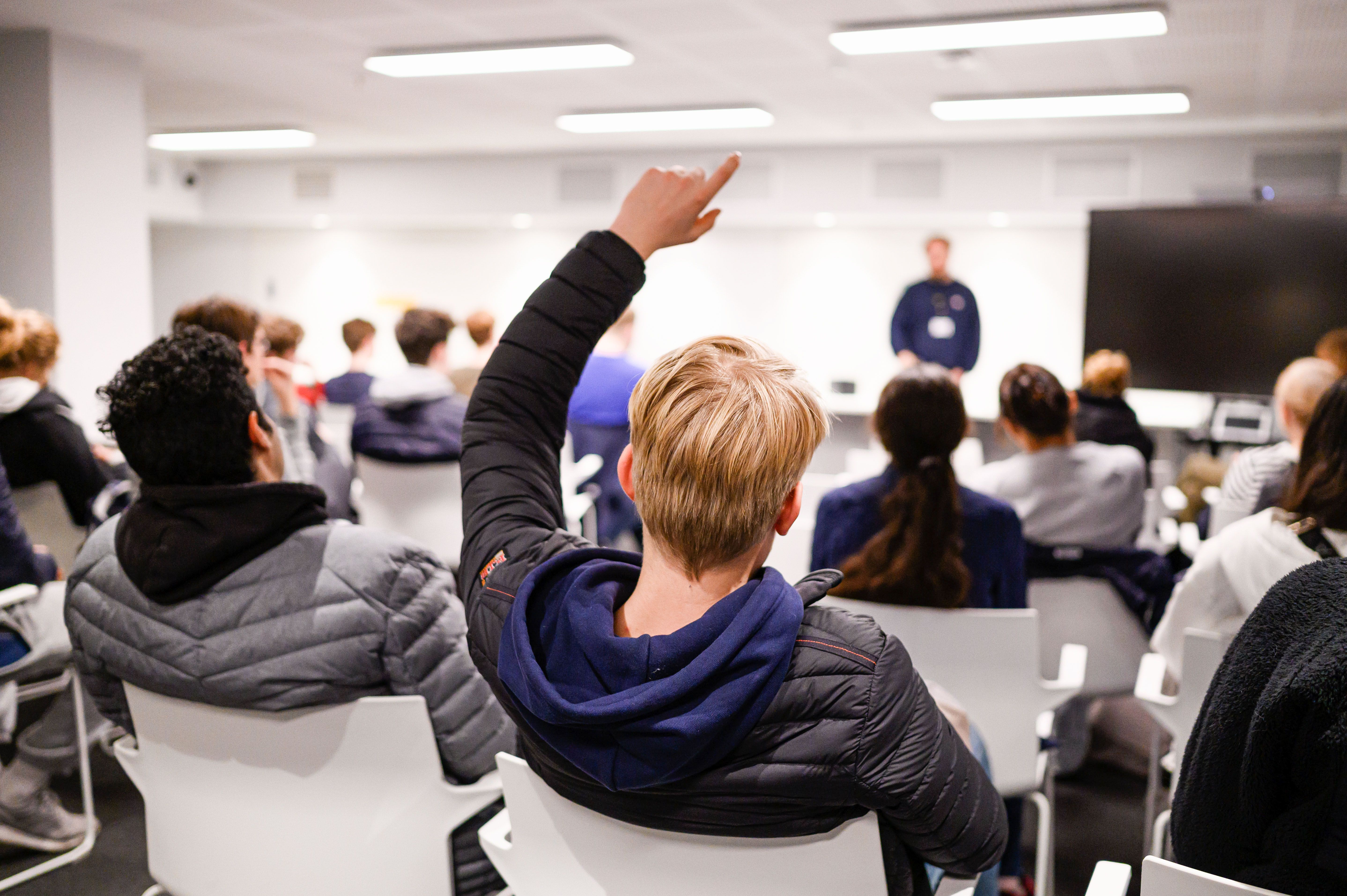 To elever sittende med ryggen til og seende mot resten av klassen og lærer. En gutt rekker opp hånden.  