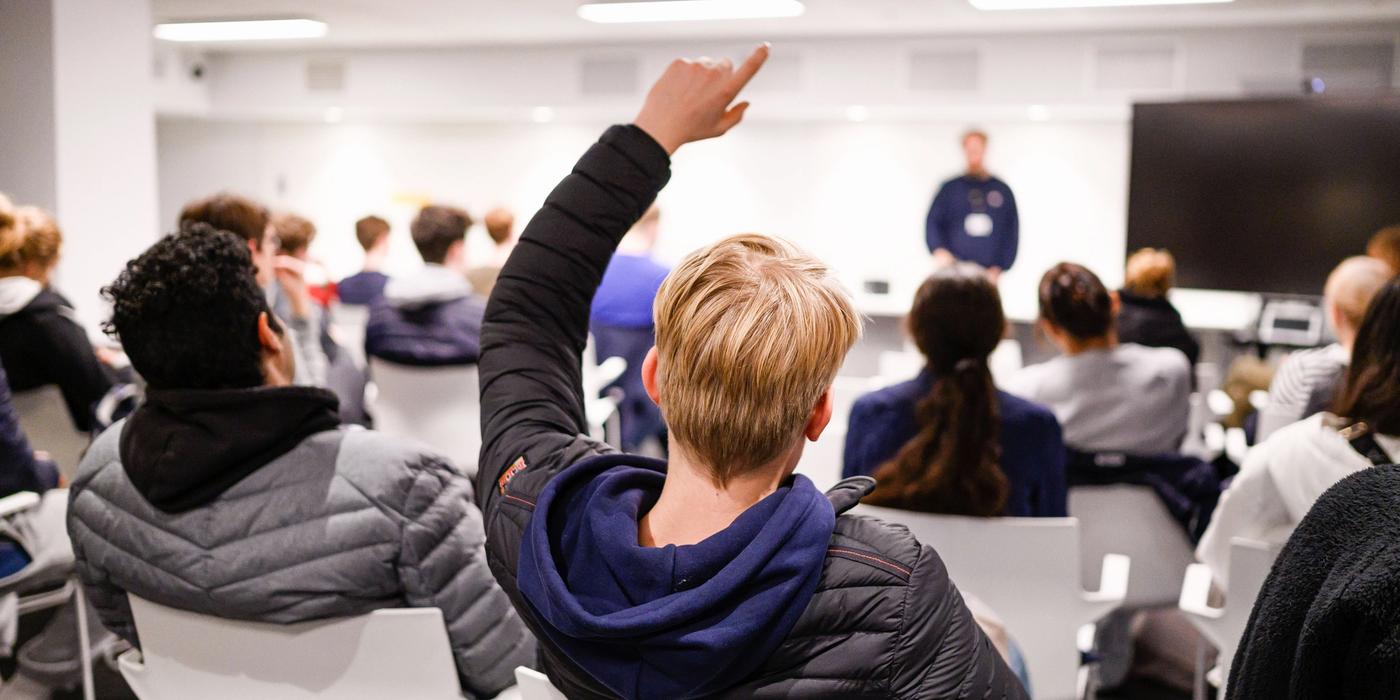 To elever sittende med ryggen til og seende mot resten av klassen og lærer. En gutt rekker opp hånden.