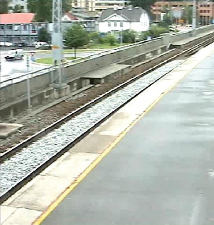 Blurred photo of a train line. On the left side og the train line there is a road where a white van passes.