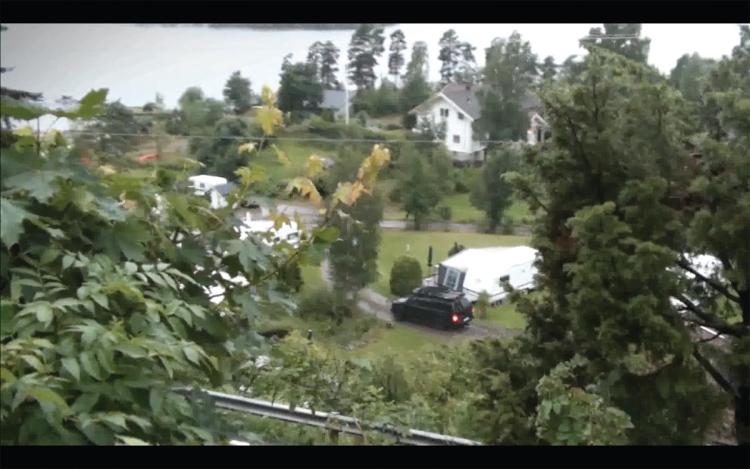 Blurry image from video recording. Landscape with trees in the front and several houses. A black car is driving along a dirt road. A lake in the background.