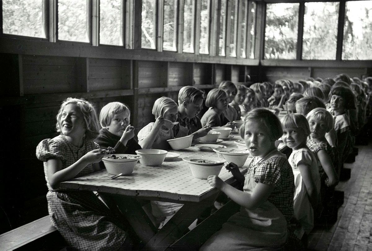 Black and white photo of a long table with children, where several eat with a spoon from a bowl. In the background a row of windows where trees can be seen.