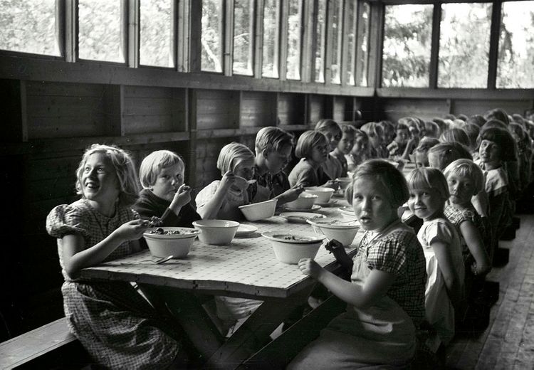 Black and white photo of a long table with children, where several eat with a spoon from a bowl. In the background a row of windows where trees can be seen.
