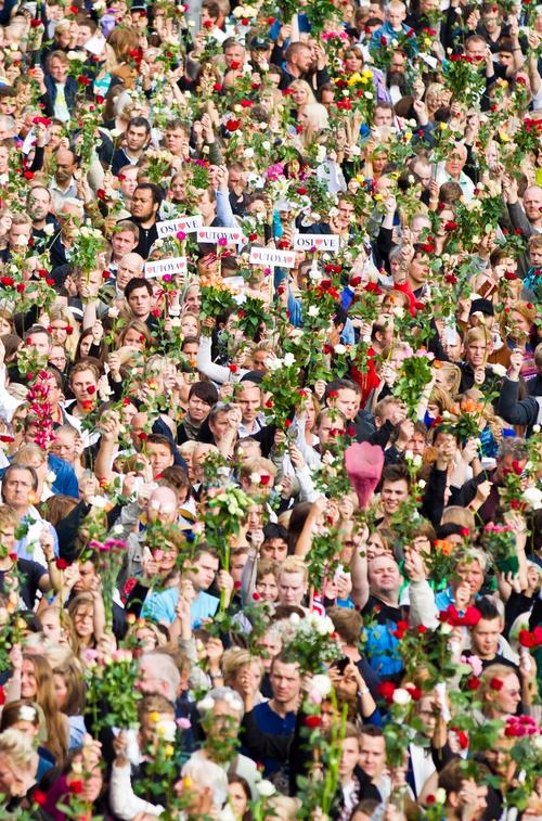 Gathering of people holding a rose in the air. Some hold placards inscribed "UTØYA" and "OSL"
