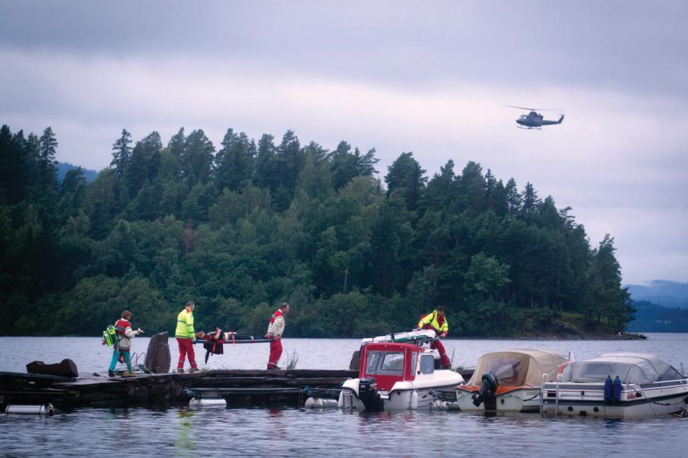 Water. Dock with three boats. Five people, where two people carry an injured person on a stretcher. Helicopter in the air. Silhouette of island with trees in background. Gray sky
