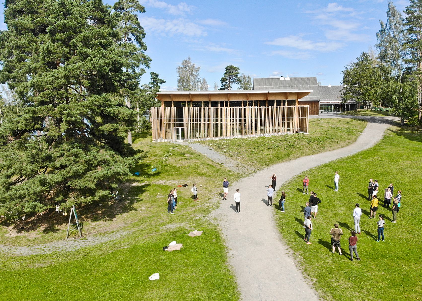 Bird's-eye view of people on a grassy area with a dirt road. In the background is a building with many wooden columns.
