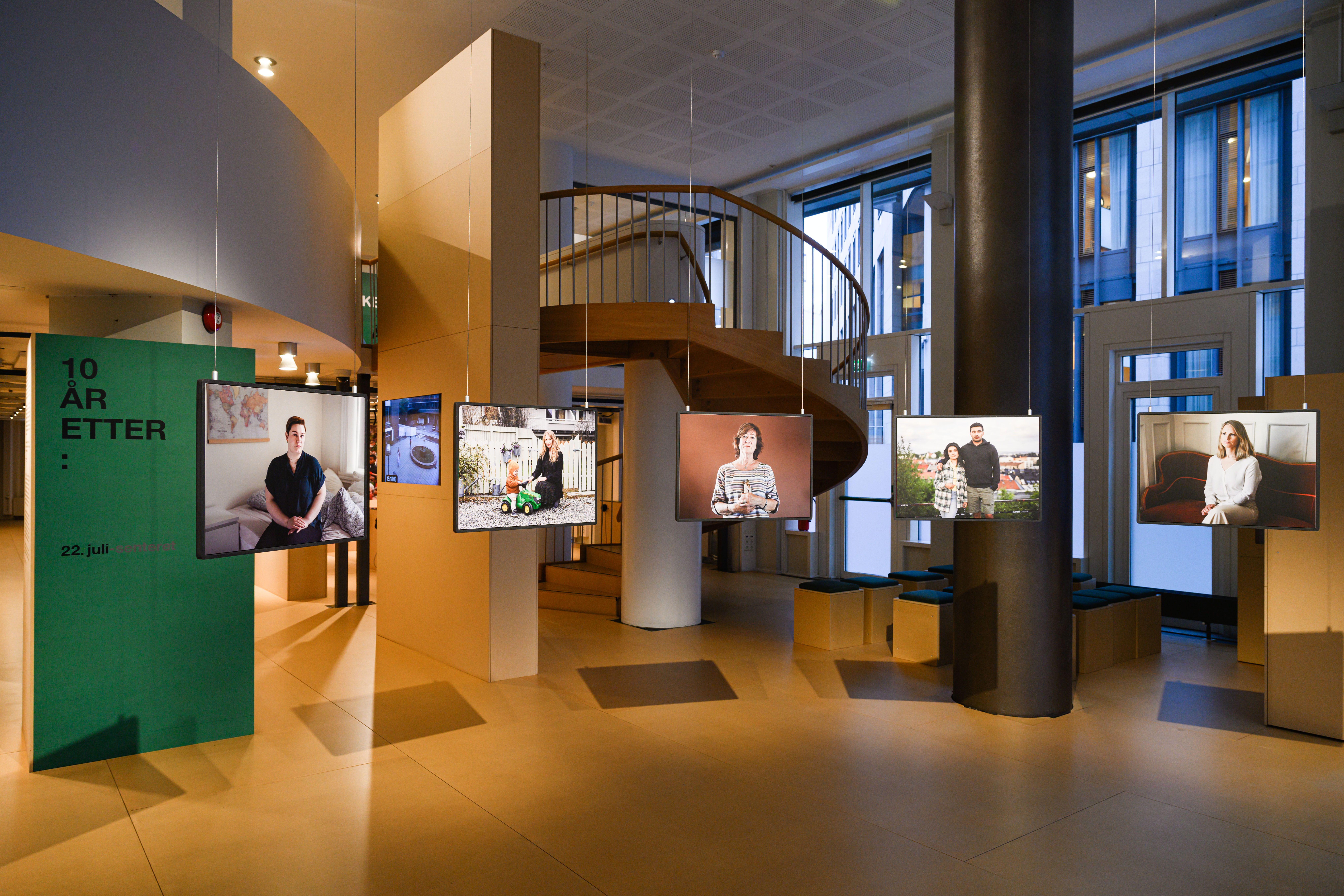 Et utstillingsrom med fem portretter som henger fra taket. Bak portrettene til venstre, en grønn plakat med skrift "10 år etter:"