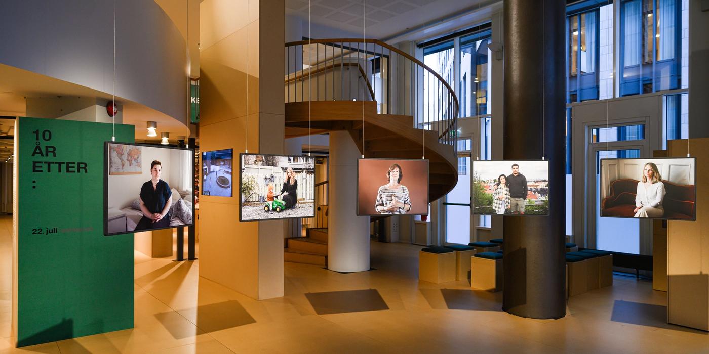 An exhibition room with five portraits hanging from the ceiling. Behind the portraits on the left, a green poster with the words "10 years after:"