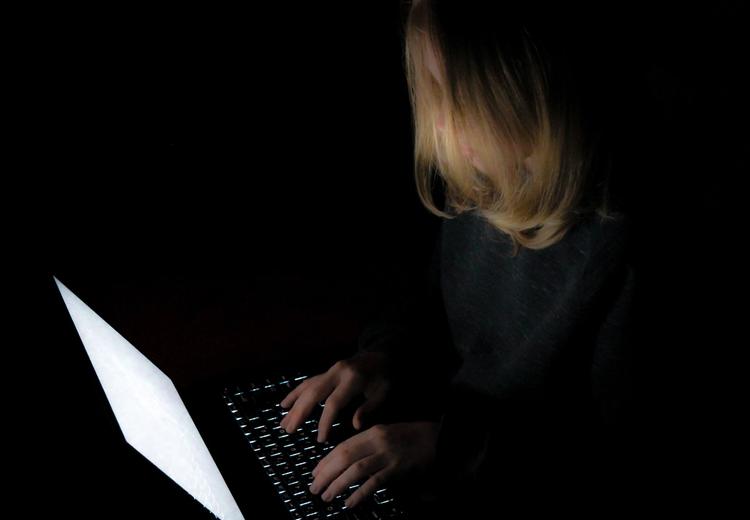 Young person with long hair with computer. The light from the PC is the only light in the room. Completely black room.