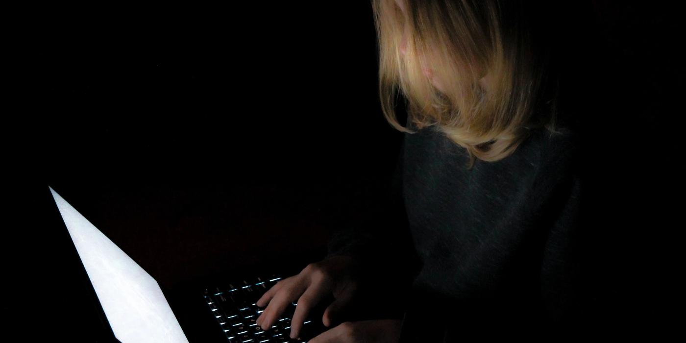 Young person with long hair with computer. The light from the PC is the only light in the room. Completely black room.