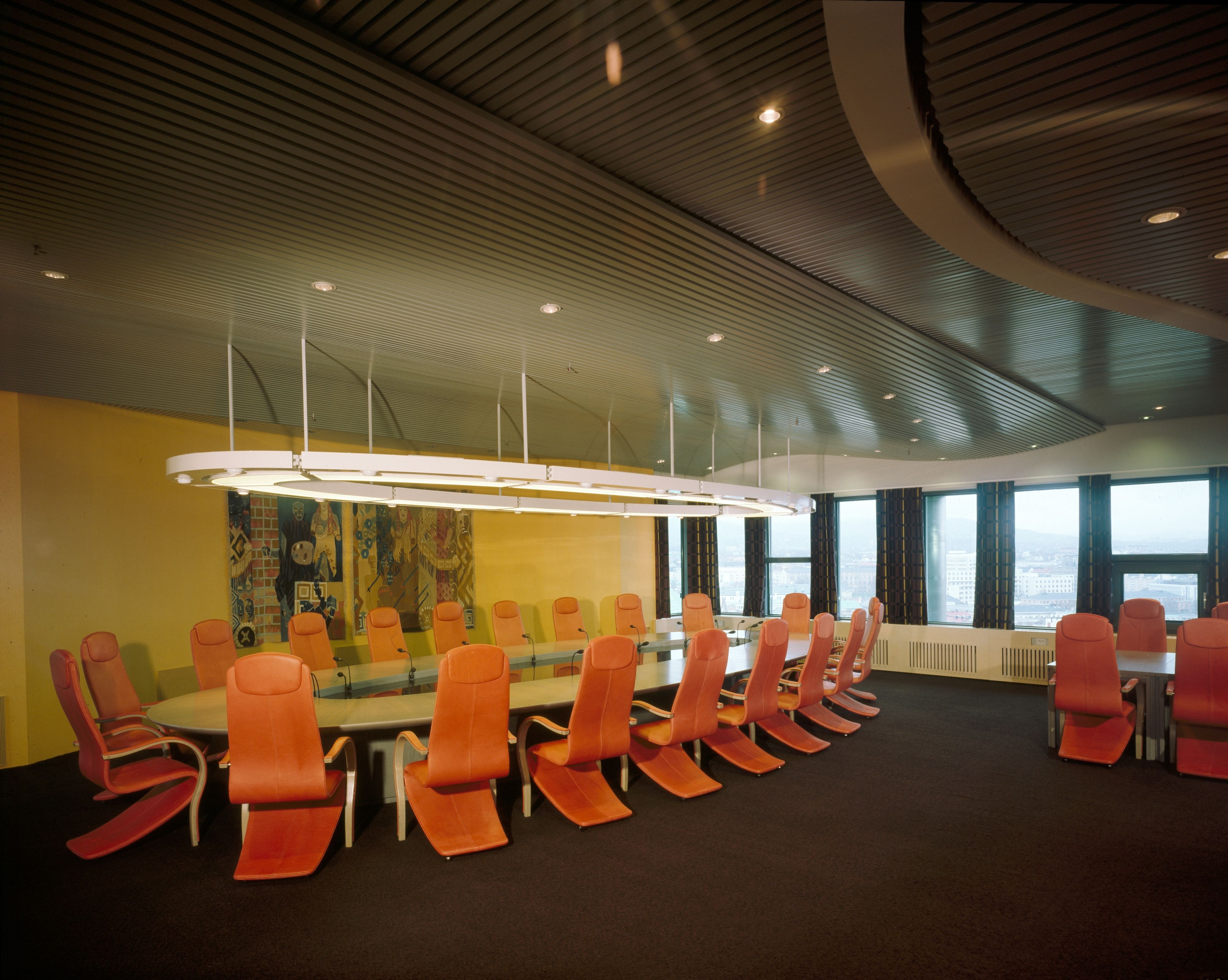 Office with oval long table with orange chairs. Yellow wall with woven pictures. In the background row of windows.