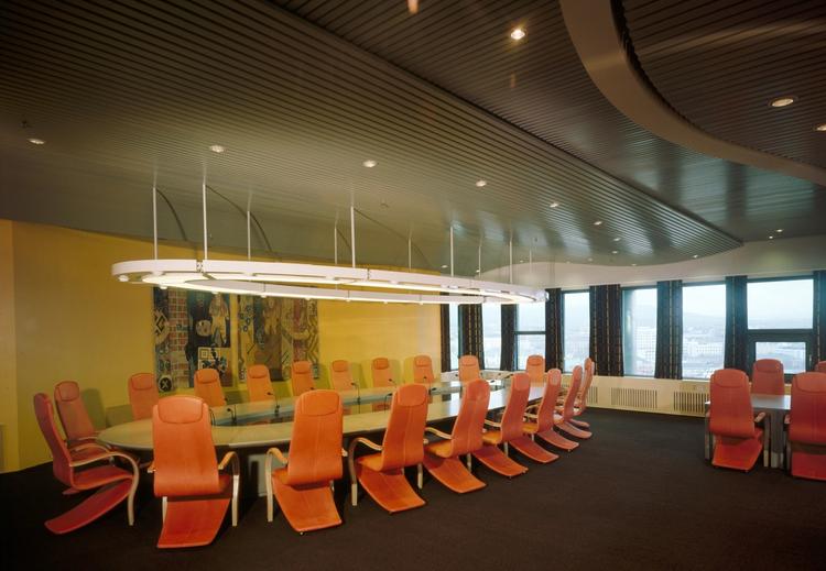 Office with oval long table with orange chairs. Yellow wall with woven pictures. In the background row of windows.