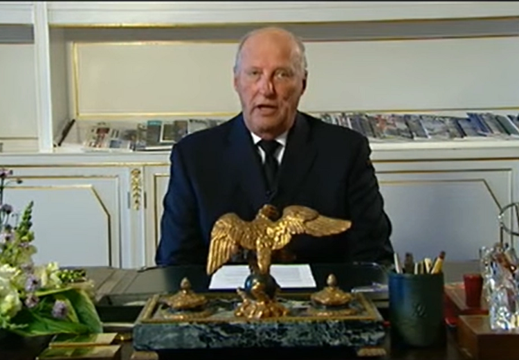 A man in a suit sits behind a desk equipped with stationery, a flower arrangement and a statue of an eagle. In the background shelf with magazines and books.