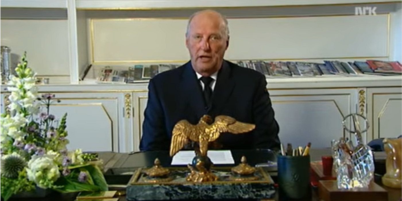 A man in a suit sits behind a desk equipped with stationery, a flower arrangement and a statue of an eagle. In the background shelf with magazines and books.