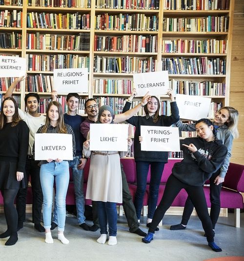 fifteen people lined up in front of bookshelf. Gray floor. Purple sofa behind the people. 10 posters with inscriptions: "Participation" "Equality" "Diversity" "Freedom of expression" "Equality" "Equality" "Tolerance" "Freedom of expression" "Freedom