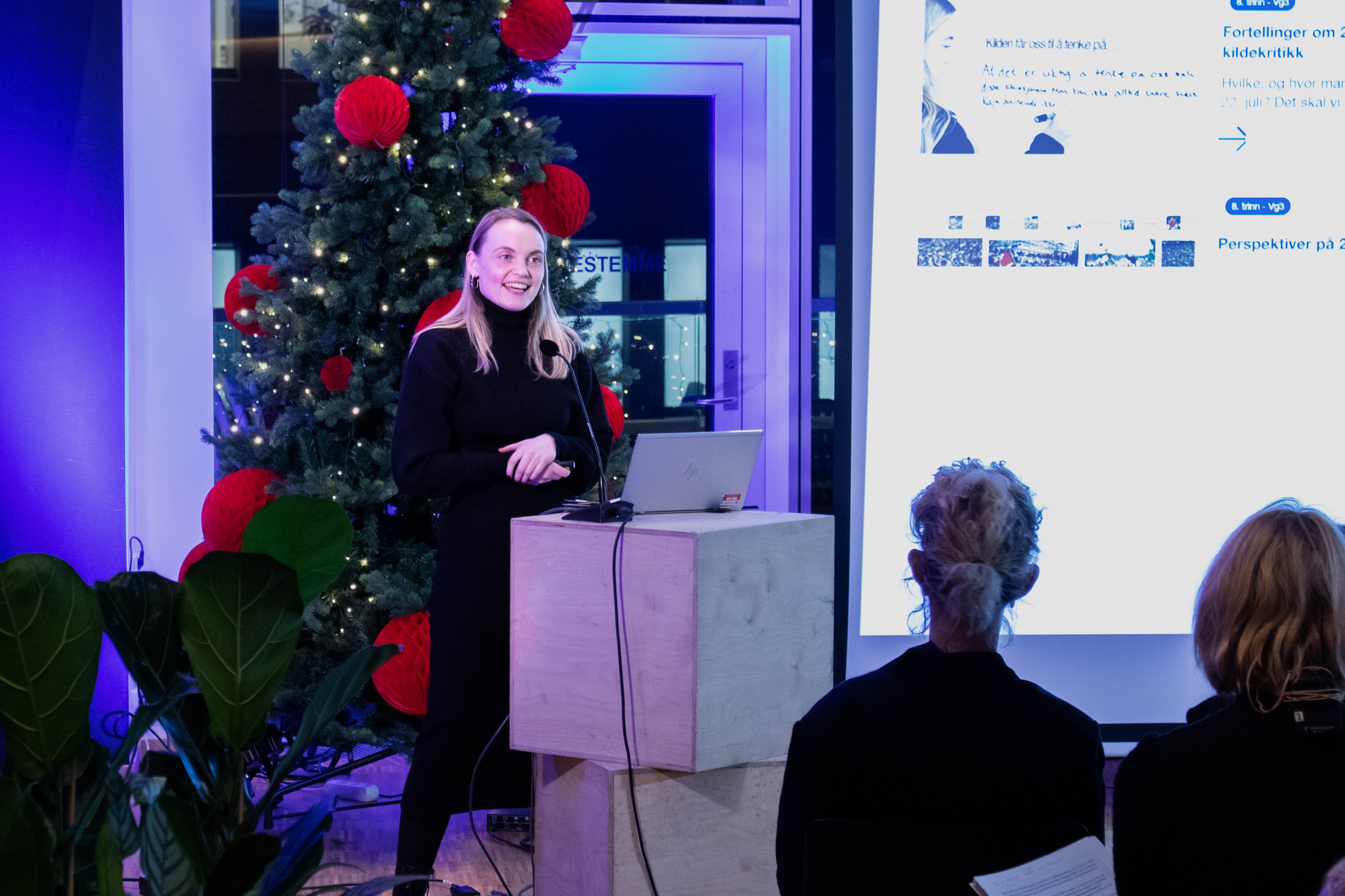 A woman stands behind a lectern and speaks to a seated audience. Behind the woman, a big screen with an indistinct projection, and a Christmas tree with large red balls.
