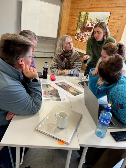 Seks mennesker sitter samlet rundt et arbeidsbord. De prater og lytter engasjert rundt noe arbeid som ligger på midten av bordet.