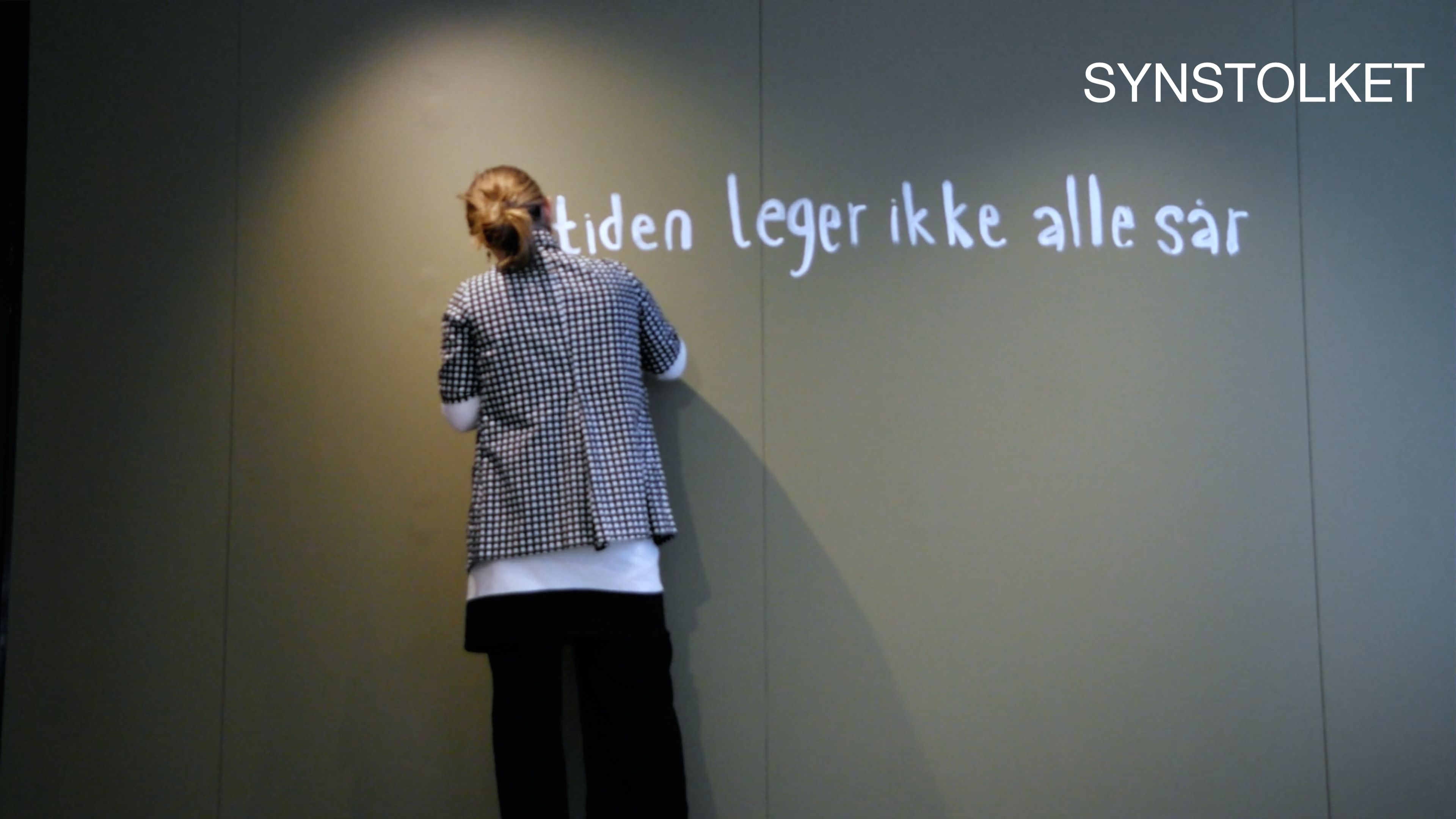 Woman, back against camera, writing with white paint on green wall. "Synstolket" (audio description) is written in the corner.