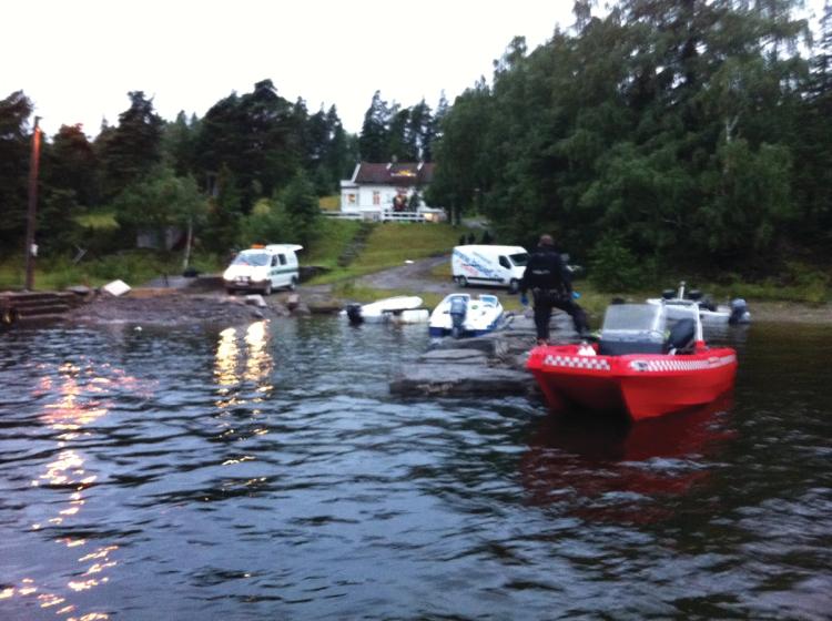Water. Three boats. A person in uniform. Two cars. Quay. White house among trees. Gray sky