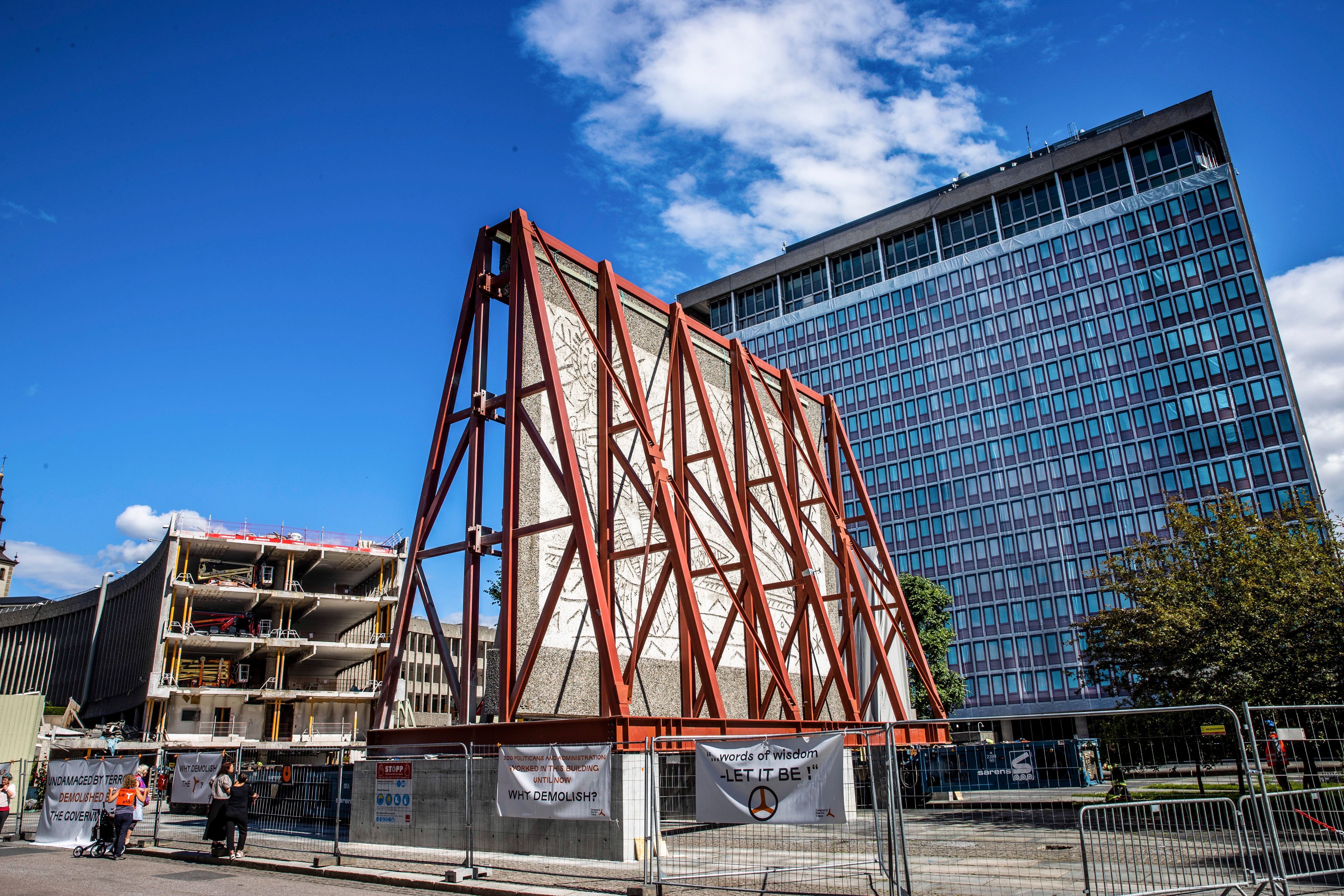 Asphalted road in the foreground. Wall section in sandblasted concrete surrounded by steel structure. Work fences and signs - More trees. Tall building with covered wall. Damaged building in the background.