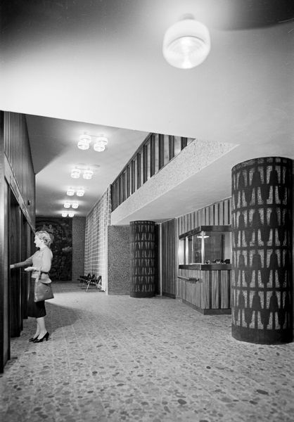 Black and white photo of a woman in an entrance hall with two ornate columns, a row of chandeliers and a reception desk.