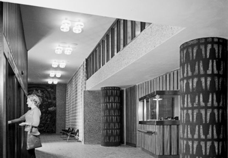 Black and white photo of a woman in an entrance hall with two ornate columns, a row of chandeliers and a reception desk.