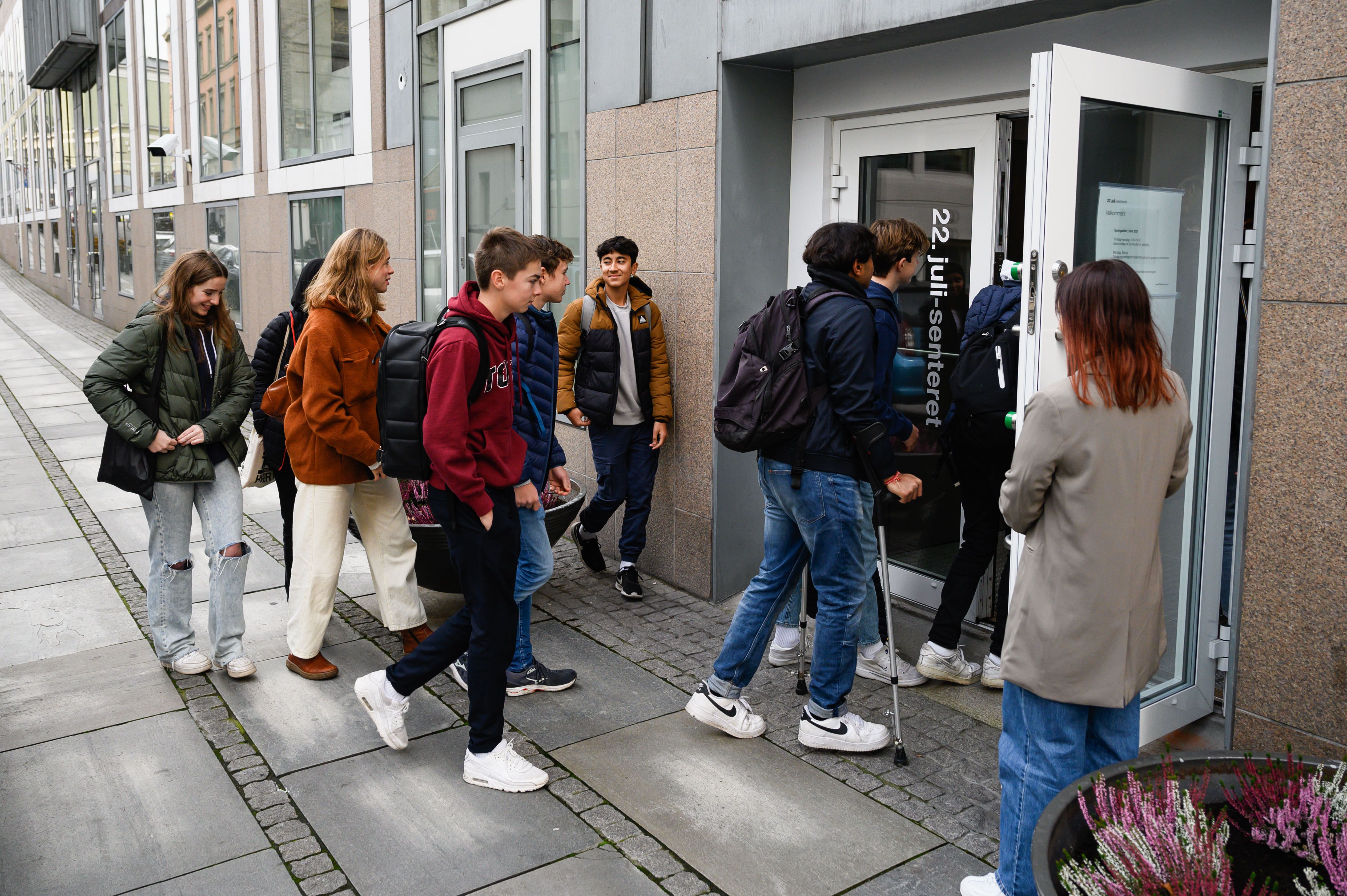 Nine young people enter the door with the logo "July 22 Center". A person holds a white door open. Building with glass windows in the background