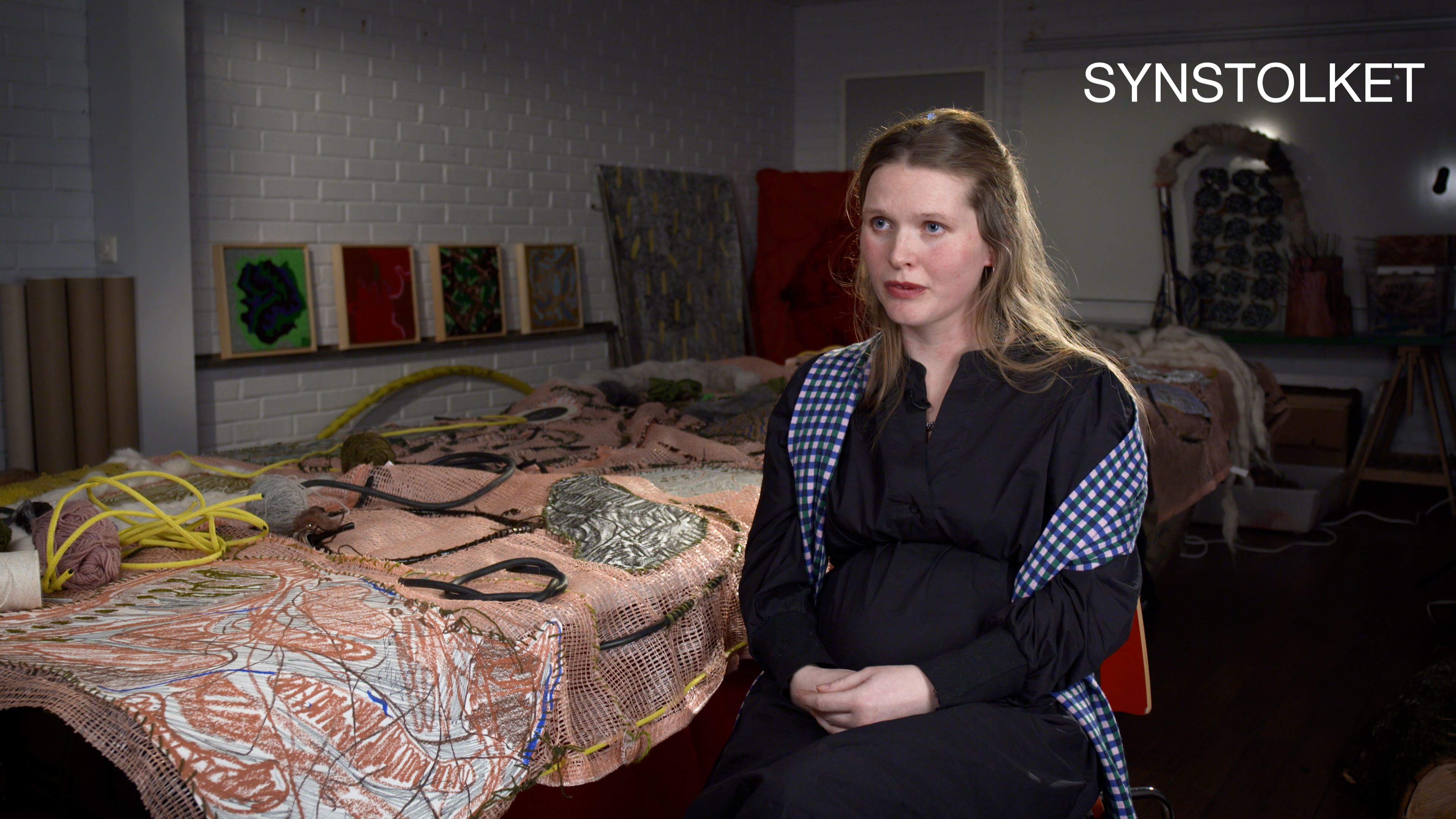 Woman in a black dress, sitting on a chair besides a large piece of artwork on a table. "Synstolket" (audio description) 