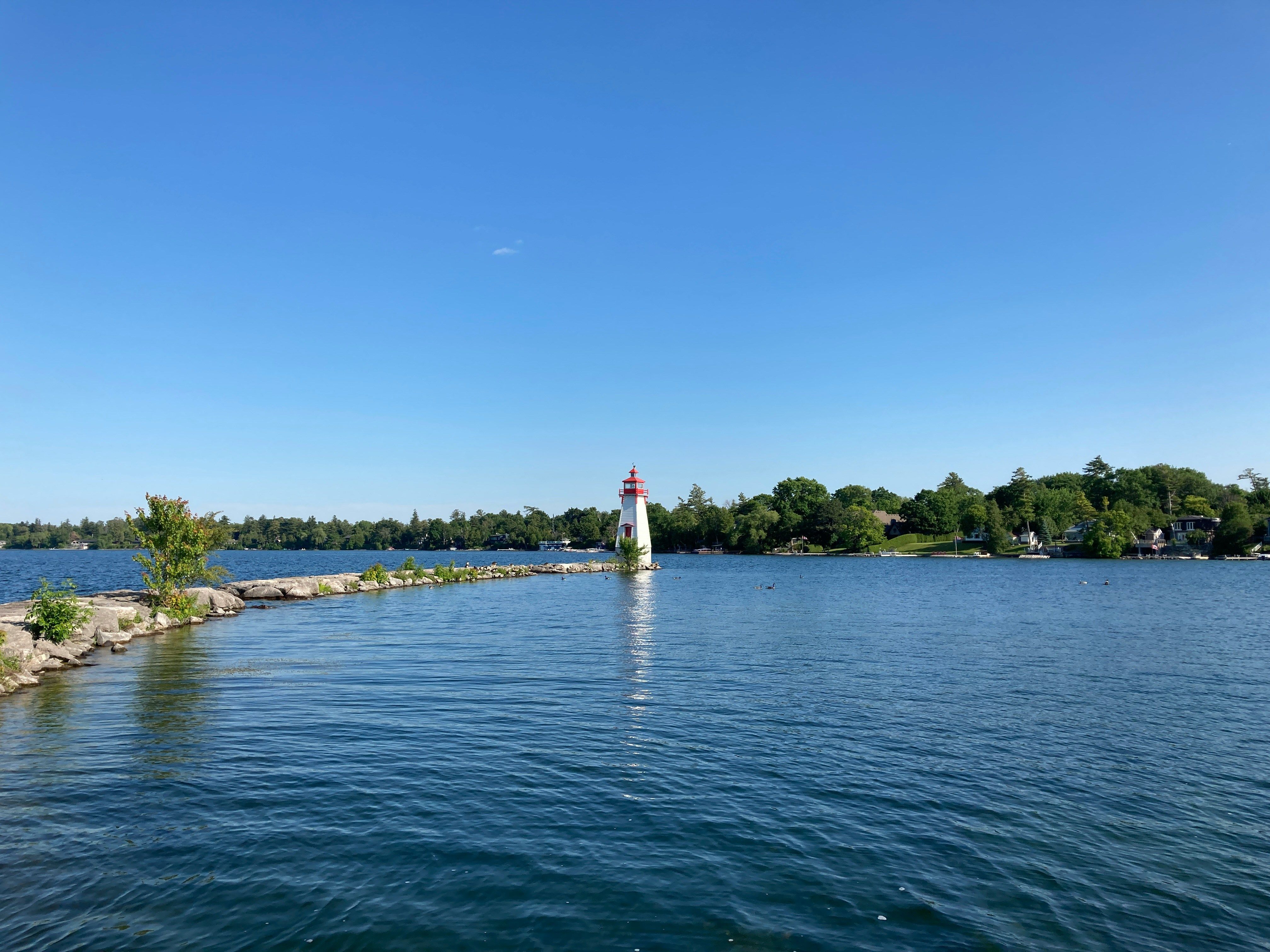White lighthouse with a red top on a rocky pier extending into a lake, with a treelined shore and clear blue sky.