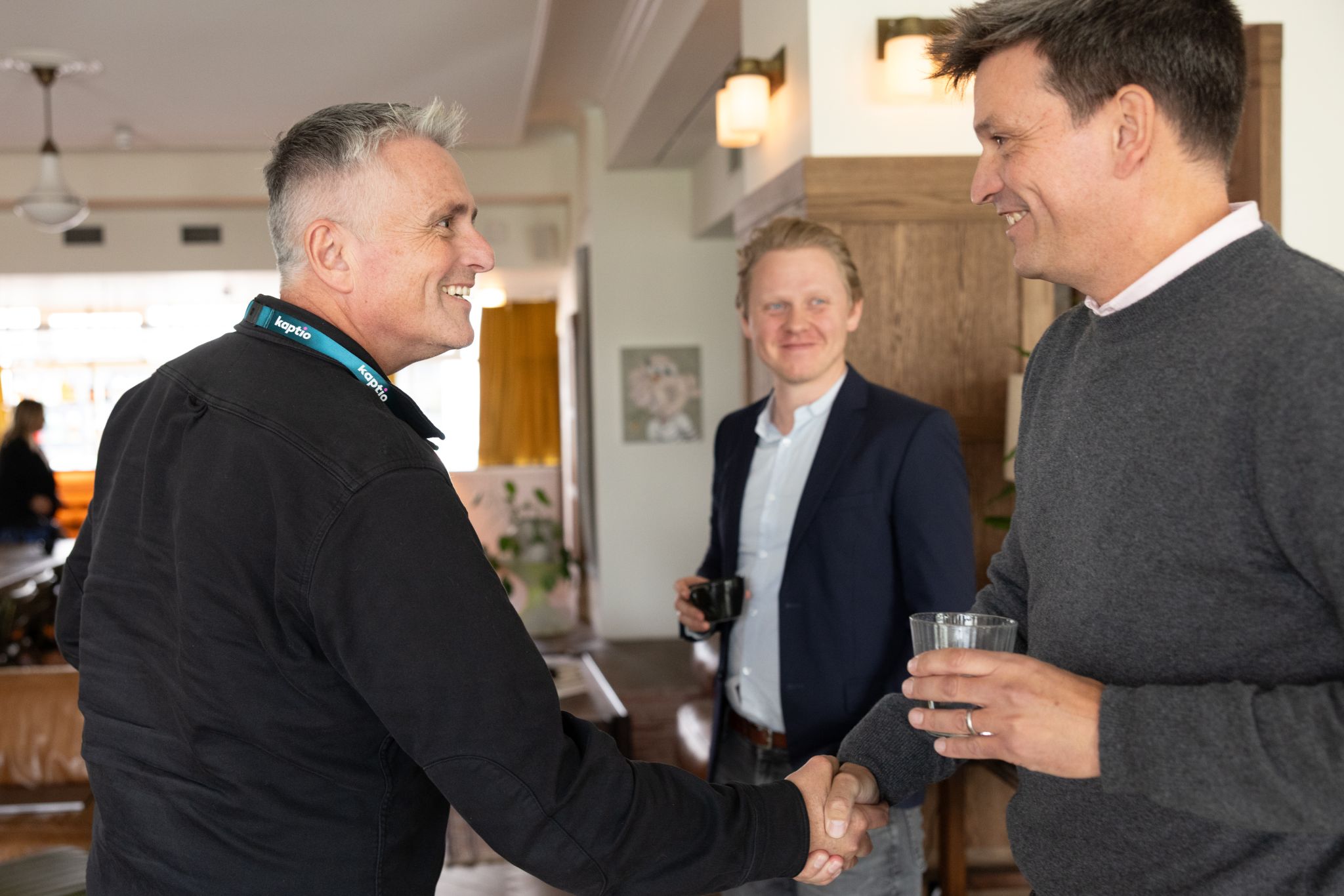 Two smiling men shaking hands, with another man smiling in the background.