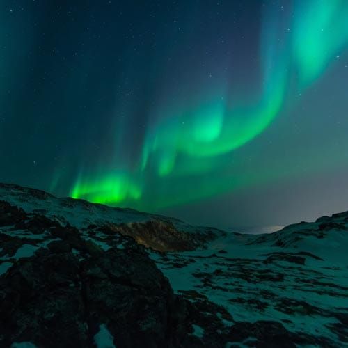 Green and blue aurora borealis in sky above snow covered rocky hills