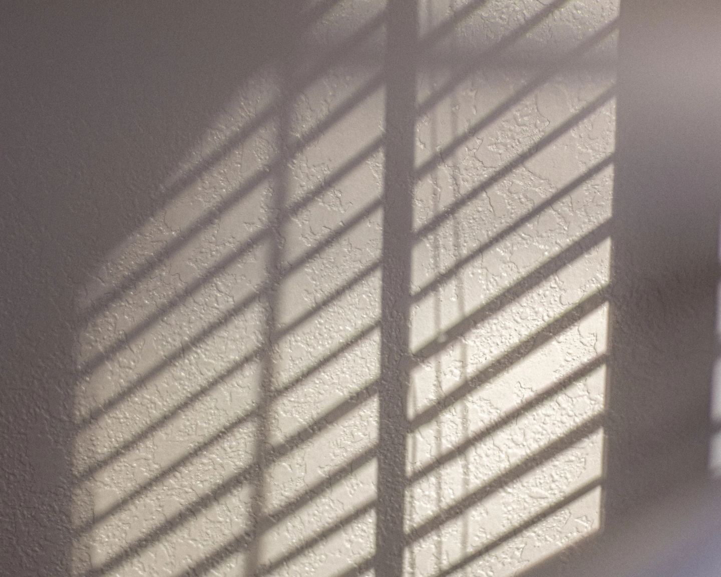 Daylight through slats casting soft banded shadows across a plain wall