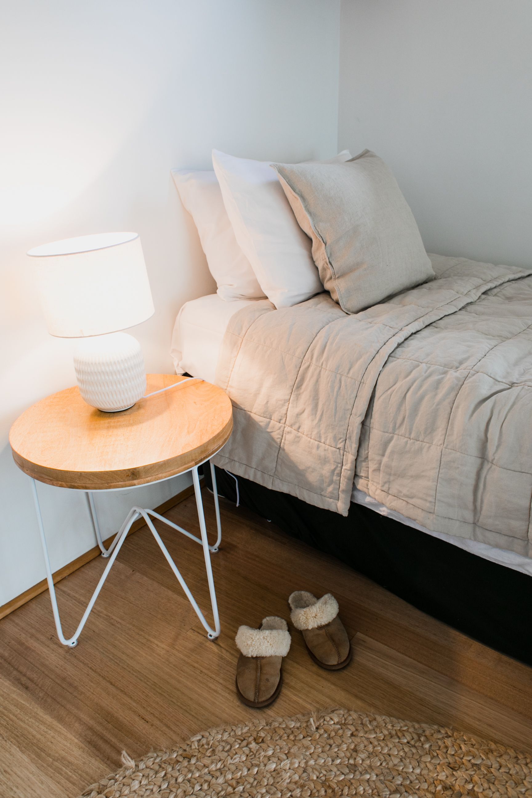 A made bed in soft neutral linen beside a timber side table and lamp