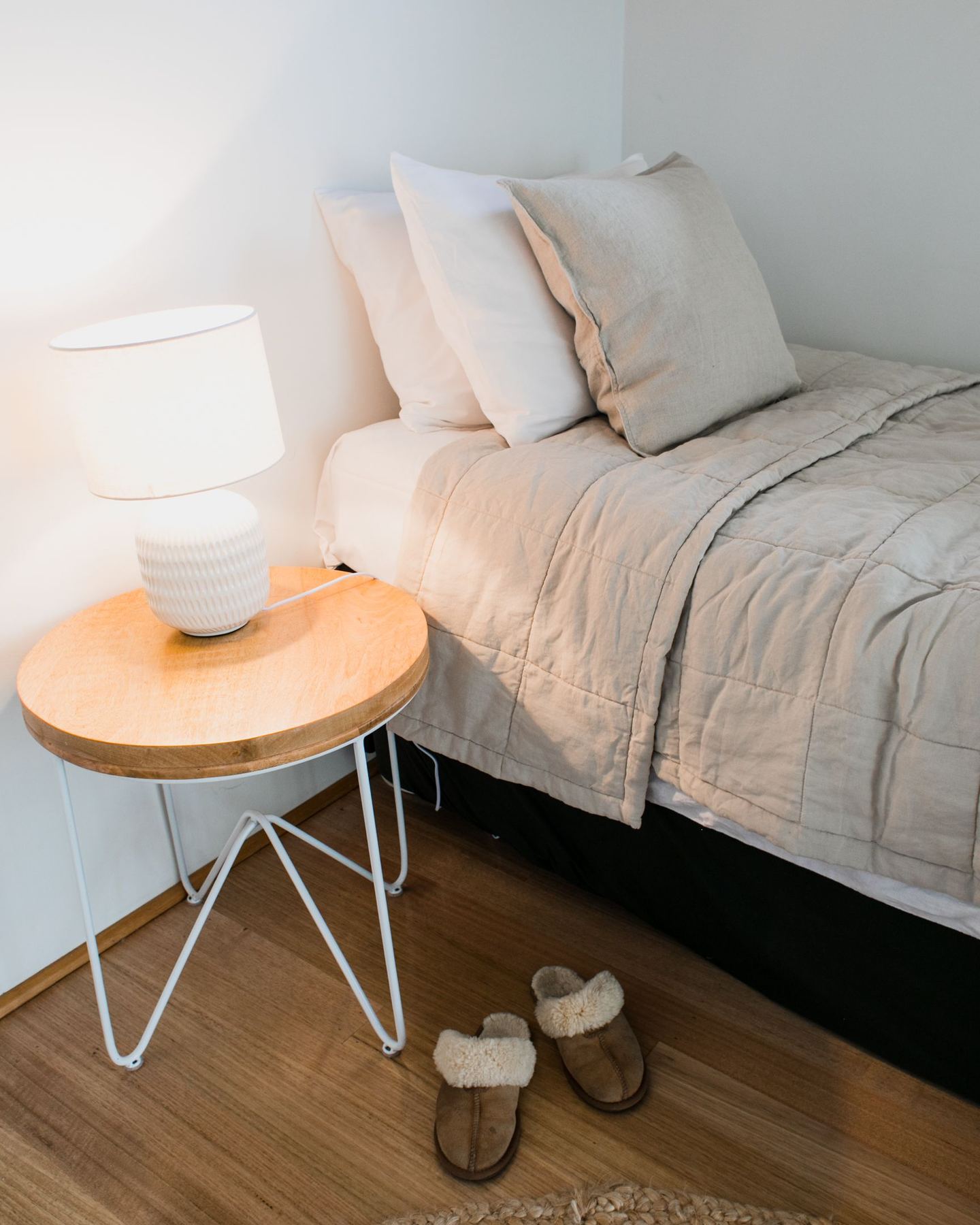 A made bed in soft neutral linen beside a timber side table and lamp