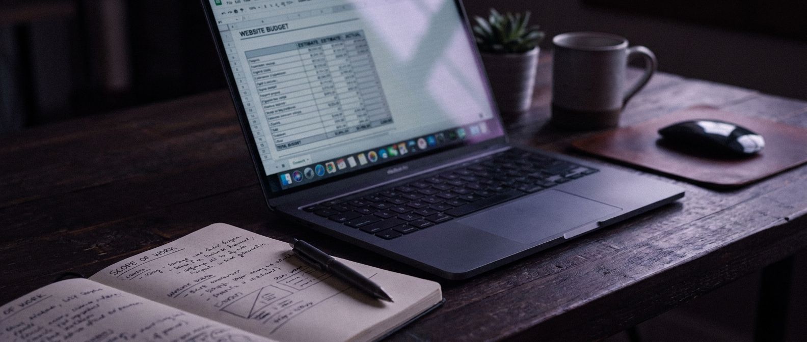 Open laptop with a small-business website budget worksheet beside a handwritten scope sheet and pen on a dark desk.