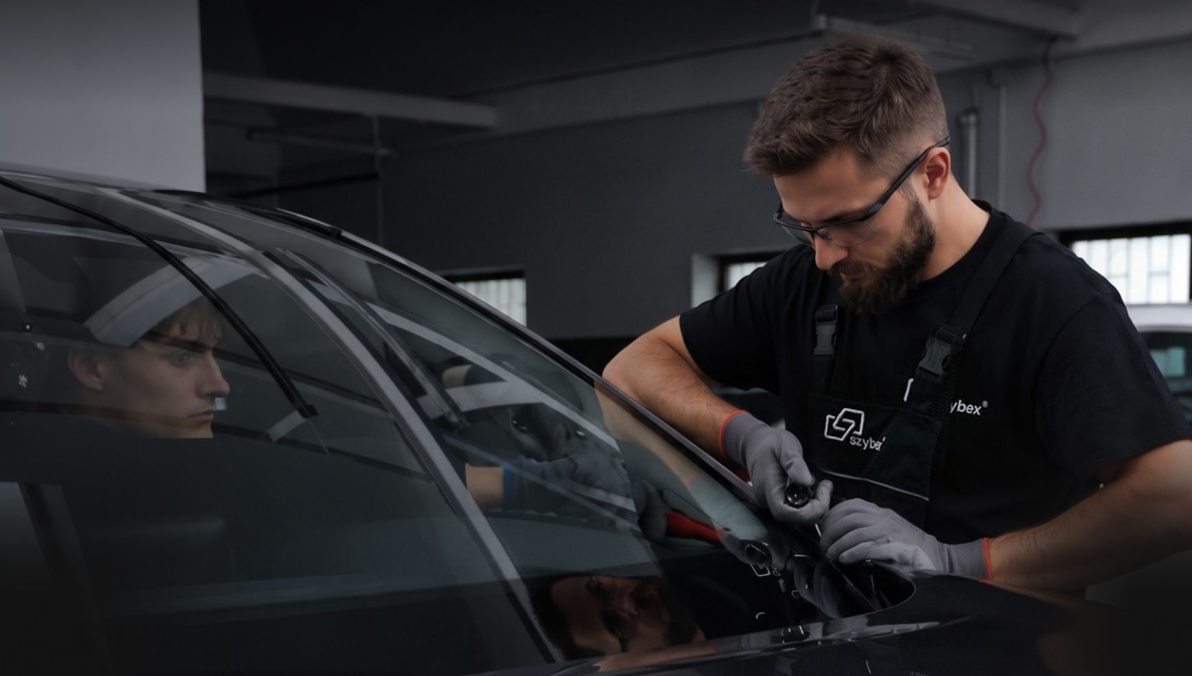 Mechanik w okularach i rękawicach pracuje przy przedniej szybie samochodu, podczas gdy w środku siedzi druga osoba.